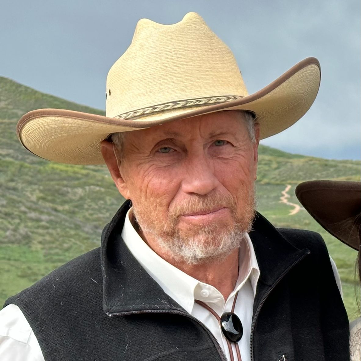 "Close-up portrait of Gregg Simonds, a man with a beard, wearing a straw cowboy hat, a white collared shirt, and a black vest. He is set against a backdrop of rolling green hills under a cloudy sky."